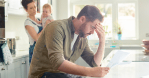 (man stressing over bills while his wife holds their baby) 