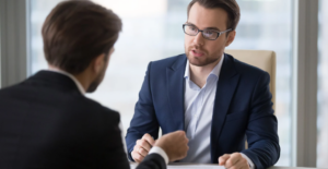 (Two professionals in a serious conversation, likely discussing finances or legal matters, across a desk in a modern office setting.)